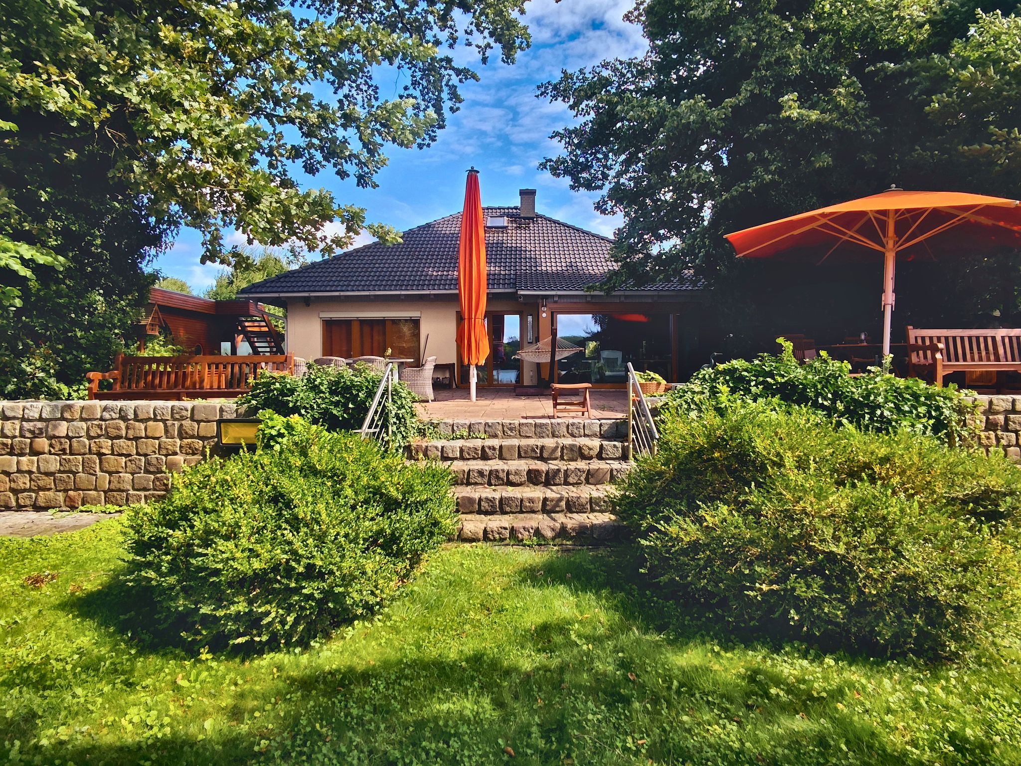 Das Einfamilienhaus in Senzig vom Garten aus: Natursteinterrasse mit Treppe, Sonnenschirme und Rasenfläche