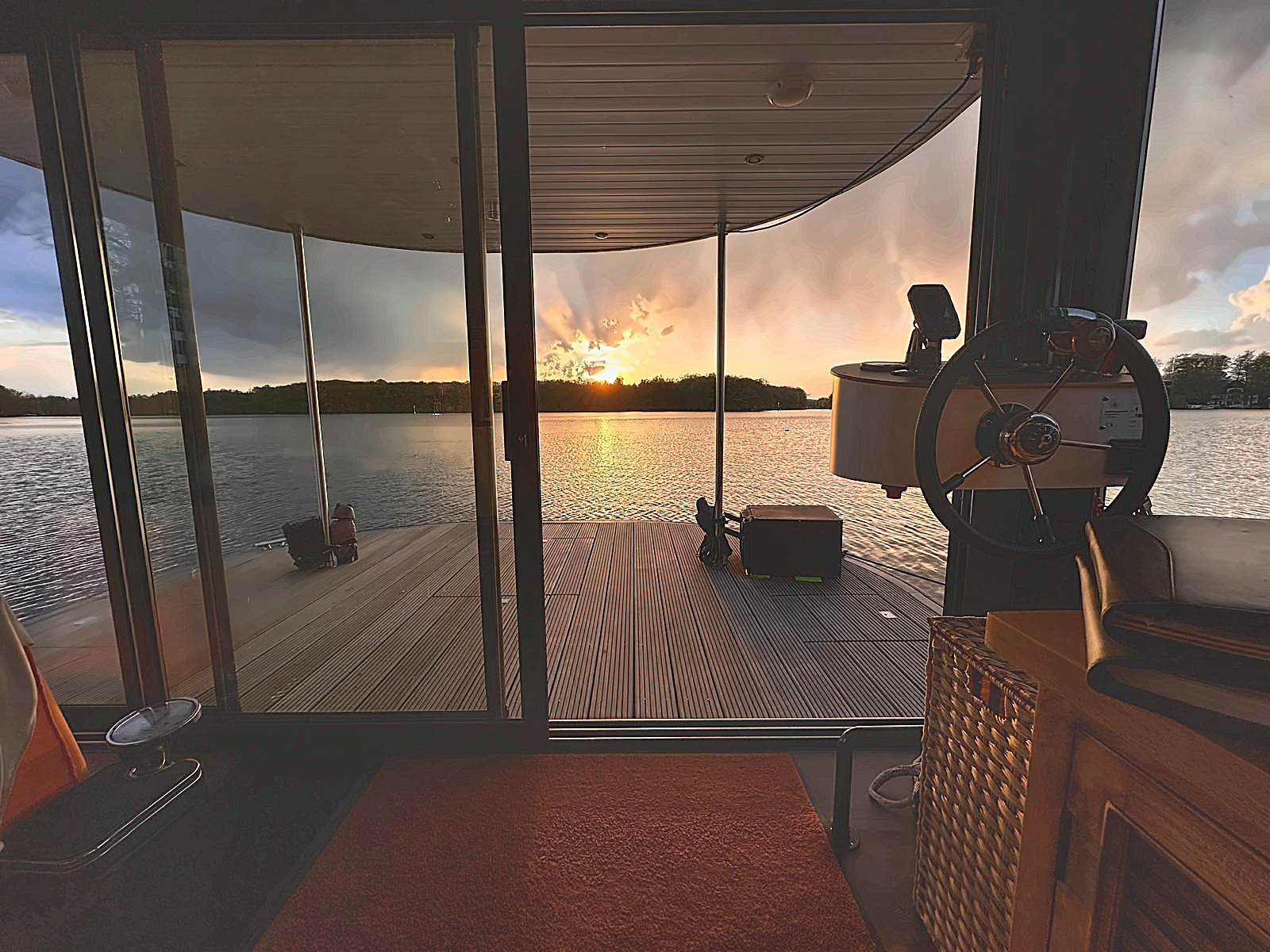 Blick aus dem Innenraum eines Hausboots auf eine Holzplattform und einen See bei Sonnenuntergang.